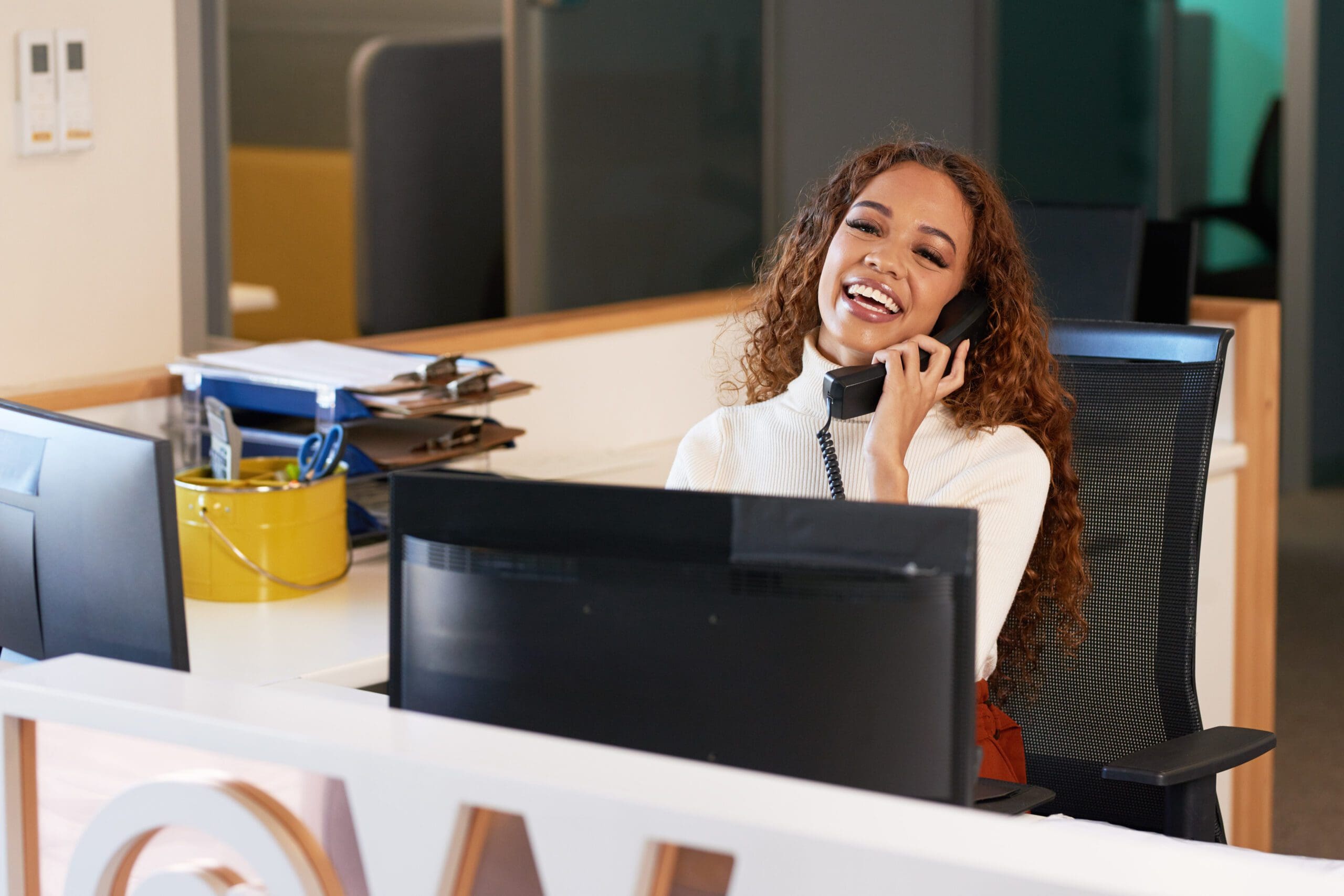 Front desk receptionist laughs on the phone, multi-ethnic woman with curly hair
