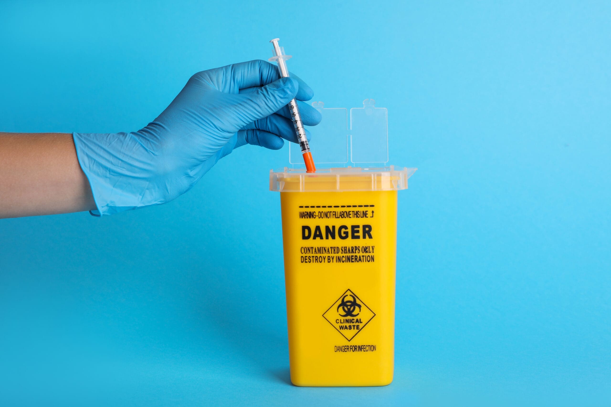 Doctor throwing used syringe into sharps container on light blue background, closeup